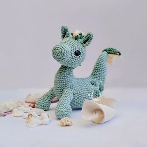 May include: A crocheted sea dragon toy with a light green body, white eyes, and a yellow crown. The toy is sitting on a white surface with seashells.