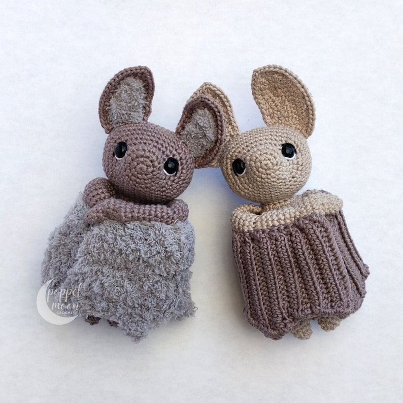 May include: Two crocheted bat plushies, one in a light brown color and the other in a gray color. Both plushies have black eyes and are wearing crocheted outfits.