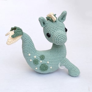 May include: A crocheted seahorse toy with a light blue body, white and green accents, and black eyes. The seahorse has a curled tail and a small fin on its back.