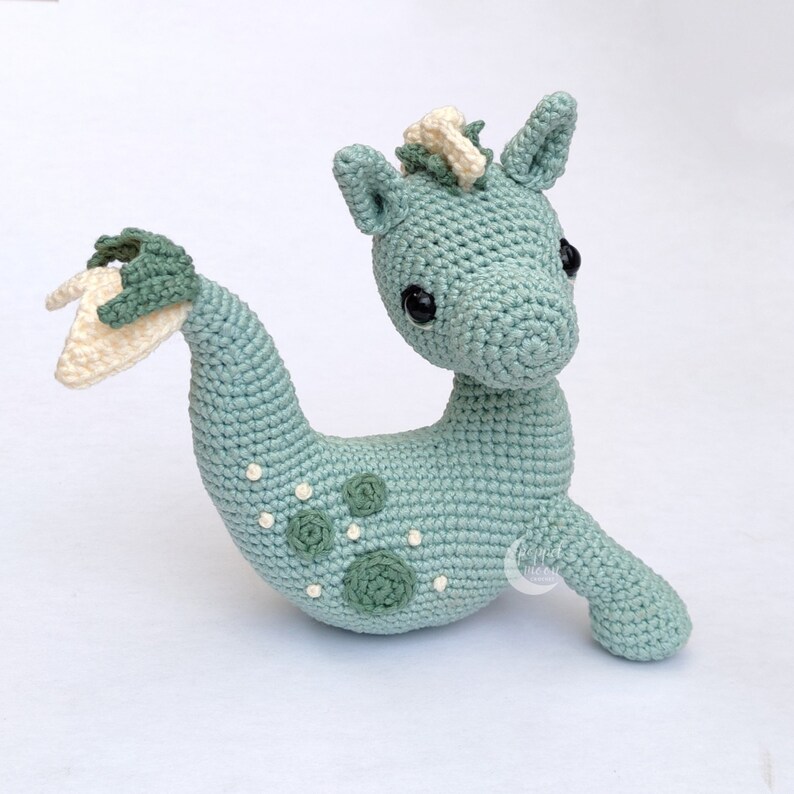 May include: A crocheted seahorse toy with a light blue body, white and green accents, and black eyes. The seahorse has a curled tail and a small fin on its back.