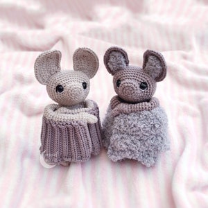 May include: Two crocheted gray bat toys with black eyes. The bat on the left is wearing a gray and white crocheted blanket. The bat on the right is wearing a gray crocheted blanket.
