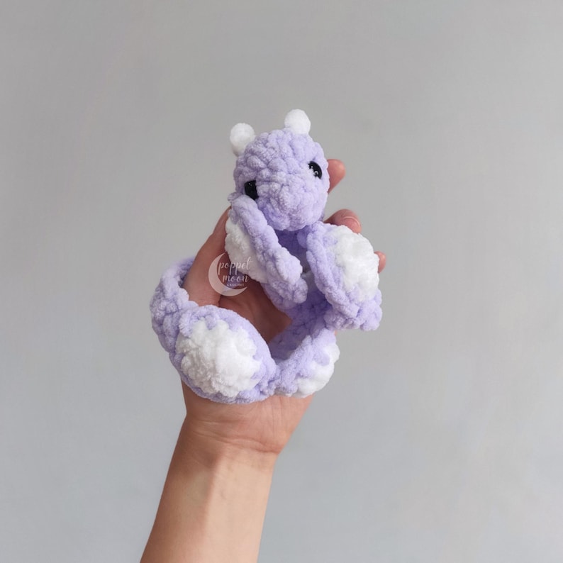 May include: A handmade, lavender crochet dragon toy with white accents. The dragon has small white horns, black eyes, and white patches on its wings and tail. The dragon is soft and plush, and appears to be held in a hand.