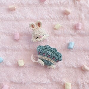 May include: A crocheted bunny hat in cream with pink accents and a crocheted turtle in green with pink accents on a pink fuzzy background.