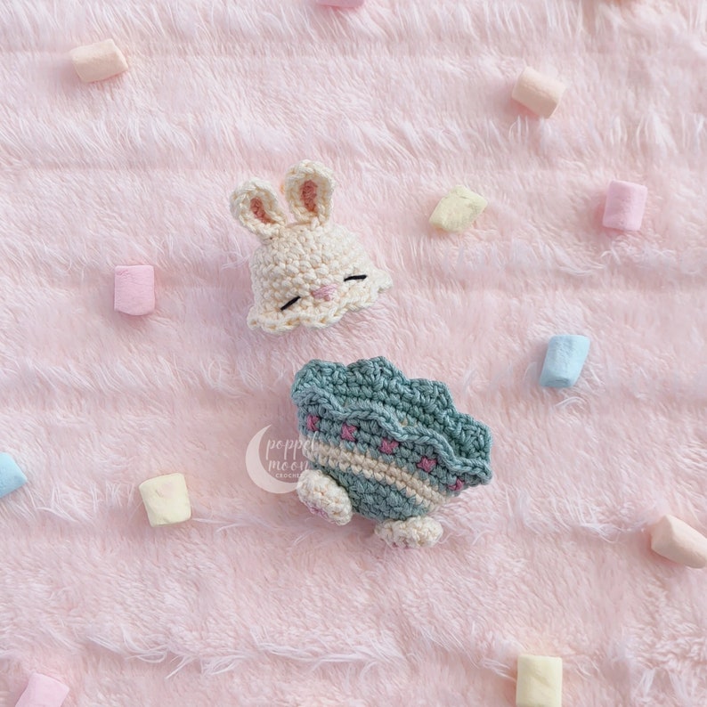 May include: A crocheted bunny hat in cream with pink accents and a crocheted turtle in green with pink accents on a pink fuzzy background.