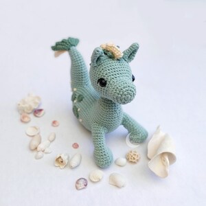 May include: A crocheted sea dragon toy with a light green body, white accents, and black eyes. The toy is surrounded by seashells on a white background.