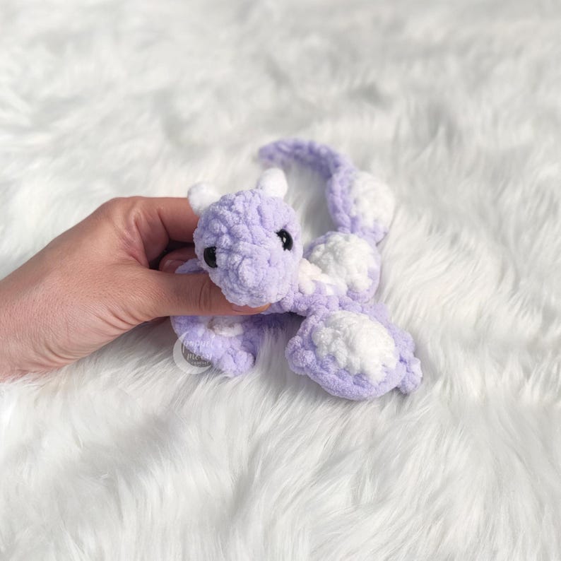 May include: A handmade, lavender and white crochet dragon toy. The dragon has a soft, textured appearance with white accents on its wings and tail. The dragon has small black eyes and white horns.