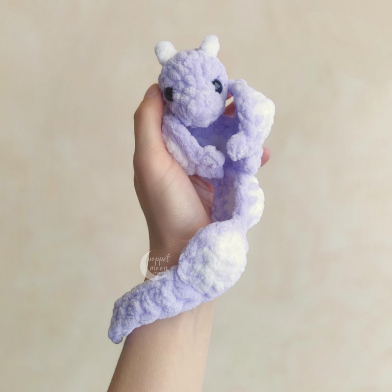 May include: A handmade, lavender-colored crochet dragon with white accents. The dragon has small white horns, black eyes, and a long tail. The plush toy is held in a hand against a neutral background.