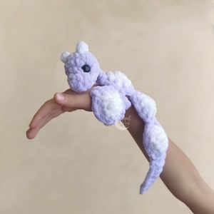 May include: A handmade, lavender and white crochet dragon toy. The dragon has a small head with two white horns, a long body, and white accents on its feet and tail. The toy is soft and plush.