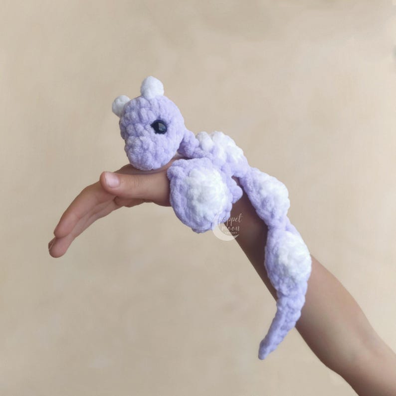 May include: A handmade, lavender and white crochet dragon toy. The dragon has a small head with two white horns, a long body, and white accents on its feet and tail. The toy is soft and plush.