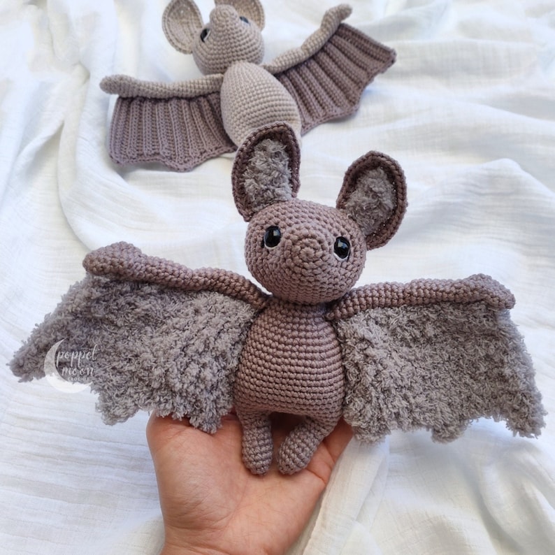 May include: A crocheted purple bat with fuzzy wings. The bat has black eyes and a small, pointed nose.