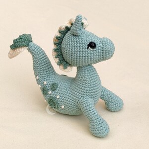 May include: A crocheted seahorse toy with a light blue body, white and green mane, and black eyes. The seahorse has a green tail and white spots on its body.
