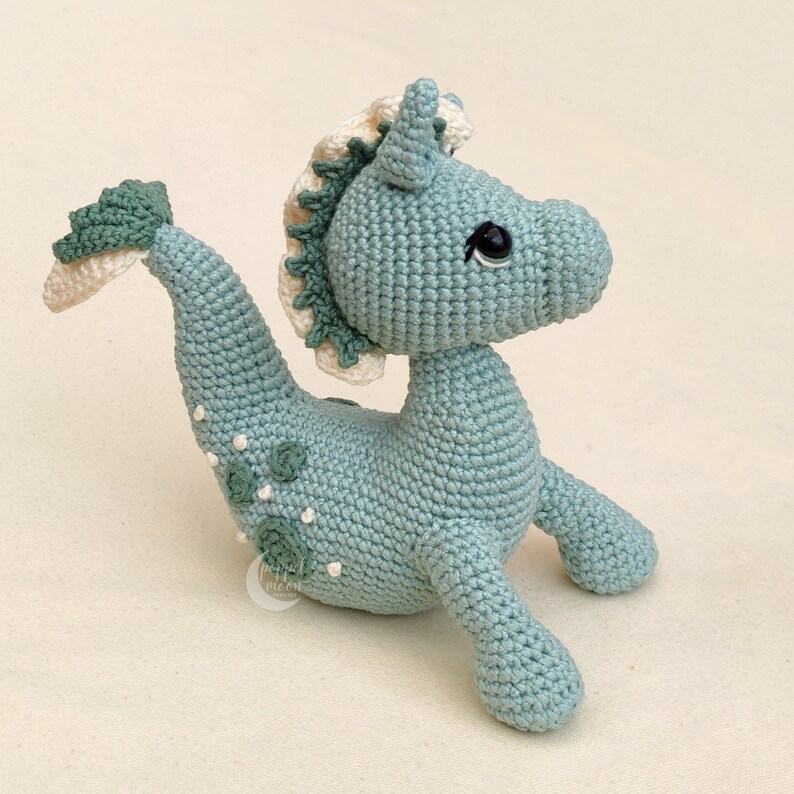May include: A crocheted seahorse toy with a light blue body, white and green mane, and black eyes. The seahorse has a green tail and white spots on its body.