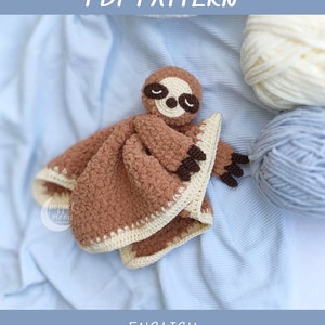 May include: A crocheted brown sloth lovey with a cream border. The sloth has brown eyes and a closed mouth. The lovey is lying on a blue blanket with white yarn and blue yarn in the background. The text "Sleepy Sloth Lovey PDF PATTERN" is at the top of the image. The text "ENGLISH Skill Level: Intermediate" is at the bottom of the image.