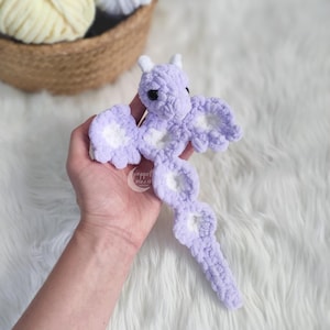 May include: A handmade, lavender crochet dragon with white accents on the wings and body. The dragon has small white horns and black safety eyes. The dragon is made of soft, plush yarn.