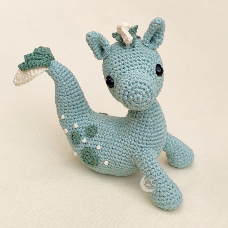 May include: A crocheted seahorse toy with a light blue body, white and green accents, and black eyes. The seahorse has a curled tail and is sitting on a white surface.