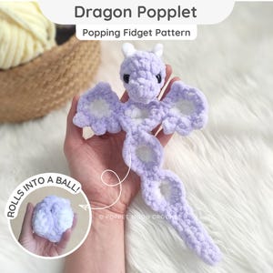 Puede incluir: Un juguete fidget de dragón tejido a ganchillo en tonos lavanda y blanco. El dragón tiene cabeza, alas y cola. El texto "Dragon Popplet" y "Popping Fidget Pattern" están en la parte superior. Una imagen más pequeña muestra el dragón enrollado en una bola.