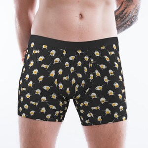 Könnte beinhalten: Schwarze Boxershorts mit einem sich wiederholenden Muster aus Cartoon-Eiern. Die Eier sind weiß mit gelben Eigelben und kleinen schwarzen Details. Der Bund ist einfarbig schwarz.