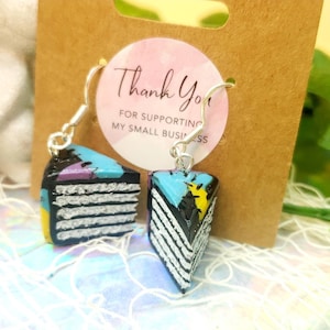 May include: A pair of dangle earrings featuring a black, white, blue, yellow, and purple cake slice design. The earrings are made of polymer clay and have silver hooks.