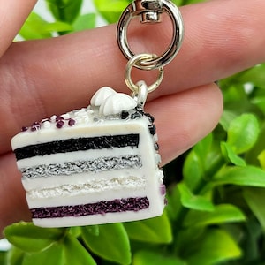May include: Asexual pride flag cake charm keychain. The charm is a slice of cake with purple, black, white, and gray layers. The cake is topped with white frosting and a few purple sprinkles.