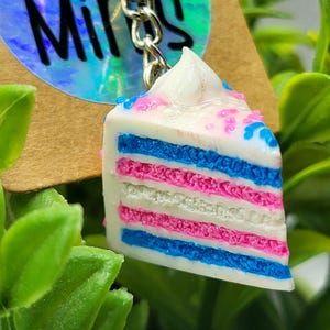 Puede incluir: Un llavero de rebanada de pastel con capas de glaseado azul, rosa y blanco. El pastel está cubierto con glaseado blanco y chispas. El llavero está sujeto a una cadena de metal plateado.