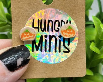 May include: A pair of pumpkin-shaped stud earrings on a brown card with the text "Hungry Minis" on a holographic sticker. The earrings have orange and white details. The card has a hole at the top.