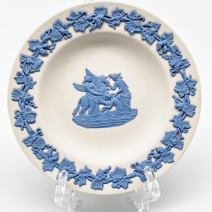 May include: A white ceramic plate with a blue and white grapevine border. The center of the plate features a blue and white relief sculpture of three figures.