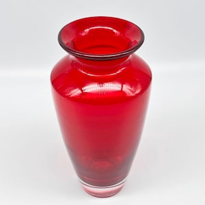 以下が含まれることがあります： フレアリムと丸みを帯びたボディが特徴的な、鮮やかな赤いガラスの花瓶。花瓶は滑らかで光沢のある仕上がりで、透明なベースが付いています。花瓶は空で、白い表面に置かれています。花瓶の高さは約25cmです。