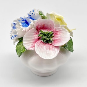 May include: A ceramic floral arrangement featuring a pink and white flower with a green center, white and blue flowers, and a yellow flower. The flowers are arranged in a white vase with a scalloped design. The arrangement is a decorative piece.