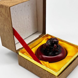 Puede incluir: Una estatua de león tallada en rojo oscuro sobre una base redonda, presentada en una caja decorativa. La caja está forrada con tela amarilla y tiene una cinta roja. La caja es marrón con un borde dorado.