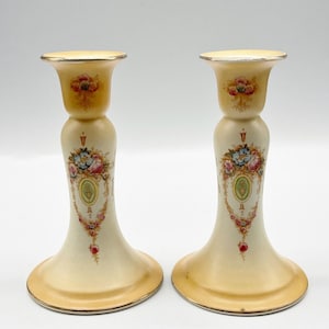 Puede incluir: Un par de candelabros de cerámica de color marfil antiguo con detalles florales y dorados. Los candelabros tienen una base en forma de campana y una parte superior cilíndrica.