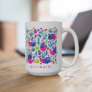 Puede incluir: Taza de cer&aacute;mica blanca con un dise&ntilde;o colorido de gatos con colas de sirena y algas marinas. El texto "purrmaids" est&aacute; impreso en rosa en la parte inferior de la taza.