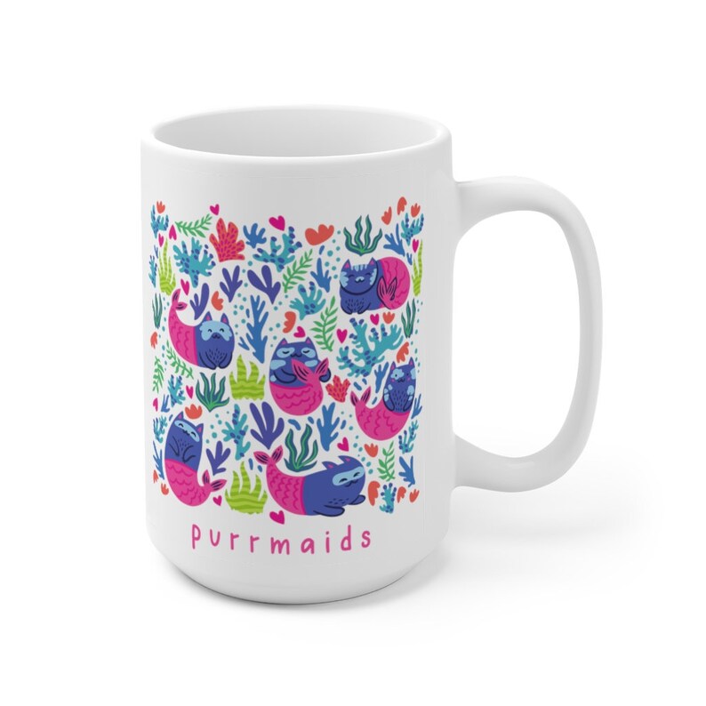 Puede incluir: Taza de cer&aacute;mica blanca con un dise&ntilde;o colorido de gatos con colas de sirena y algas marinas. El texto "purrmaids" est&aacute; impreso en rosa en la parte inferior de la taza.