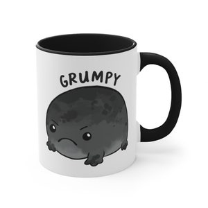May include: White ceramic coffee mug with a black rim and handle. The mug features a cartoon illustration of a grumpy black frog with the word "GRUMPY" printed above it.