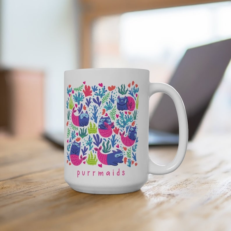 Puede incluir: Taza de cer&aacute;mica blanca con un dise&ntilde;o colorido de gatos con colas de sirena y algas marinas. El texto "purrmaids" est&aacute; impreso en rosa en la parte inferior de la taza.
