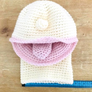 Peut inclure: Un bonnet blanc en crochet avec une bordure rose en crochet et un petit pompon blanc sur le dessus. Le bonnet est pos&eacute; sur une surface en bois avec un m&egrave;tre ruban bleu.