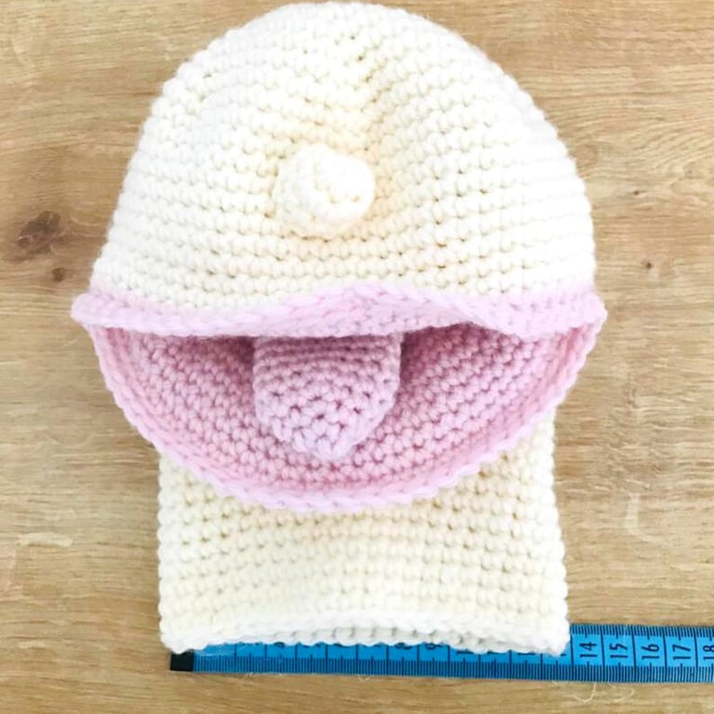 Peut inclure: Un bonnet blanc en crochet avec une bordure rose en crochet et un petit pompon blanc sur le dessus. Le bonnet est pos&eacute; sur une surface en bois avec un m&egrave;tre ruban bleu.