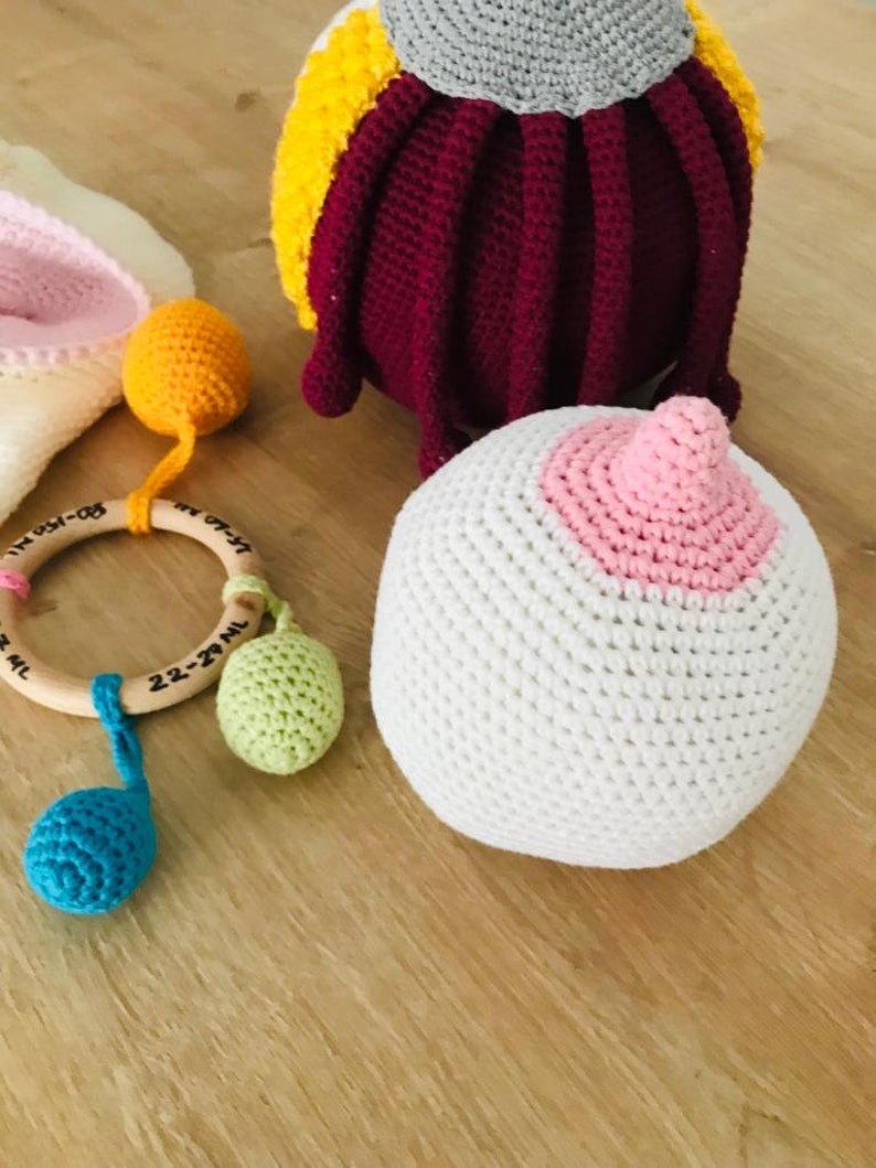 Peut inclure: Un ensemble de jouets pour b&eacute;b&eacute; en crochet, comprenant un hochet avec un anneau en bois, une balle blanche avec un cercle rose et une pieuvre multicolore. Les jouets sont sur une surface en bois.