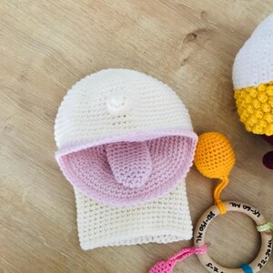 Peut inclure: Chapeau &agrave; capuche blanc en crochet avec une langue rose en crochet qui sort. Le chapeau est sur une surface en bois avec un jouet en anneau en bois avec des perles color&eacute;es.