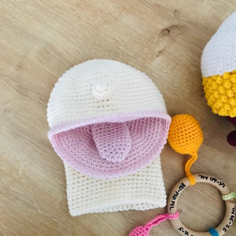 Peut inclure: Chapeau &agrave; capuche blanc en crochet avec une langue rose en crochet qui sort. Le chapeau est sur une surface en bois avec un jouet en anneau en bois avec des perles color&eacute;es.