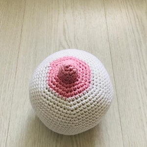 Peut inclure: Une boule blanche en crochet avec un cercle rose en crochet sur un c&ocirc;t&eacute;. Le cercle a une petite bosse rose en crochet au centre.