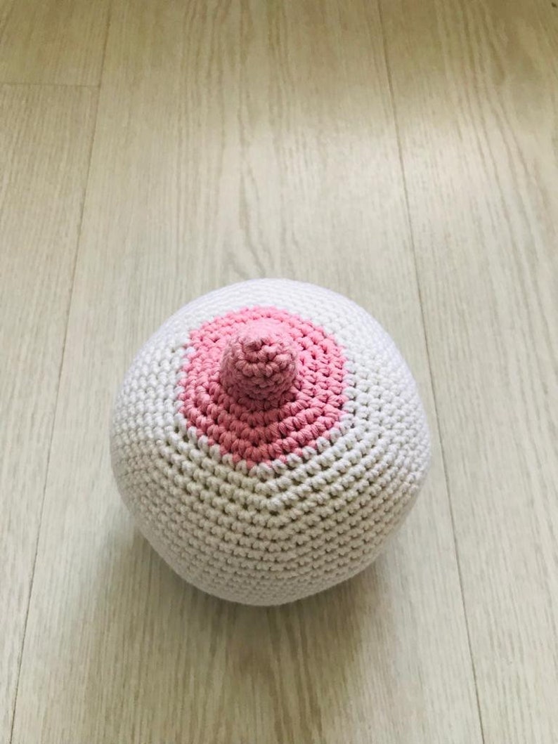 Peut inclure: Une boule blanche en crochet avec un cercle rose en crochet sur un c&ocirc;t&eacute;. Le cercle a une petite bosse rose en crochet au centre.