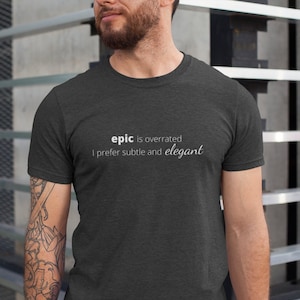 以下が含まれることがあります： 白文字で「epic is overrated I prefer subtle and elegant」と書かれた、濃いグレーの杢調Tシャツです。