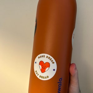 May include: A brown reusable water bottle with a white sticker that says "Make Bread Eat Bread" with a cartoon heart in the middle. The bottle has a blue "owala" logo on the bottom.