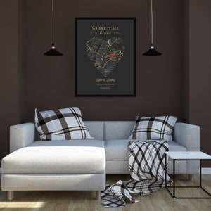 May include: A black and gold framed print with a heart-shaped map of a city. The map is outlined in gold and has a red dot in the center. The text "Where it all began" is at the top of the print, and the names "Tyler & Emma" and a date are at the bottom. The print is hanging on a brown wall above a white sectional sofa with black and white patterned pillows and a throw blanket.