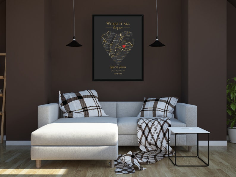May include: A black and gold framed print with a heart-shaped map of a city. The map is outlined in gold and has a red dot in the center. The text "Where it all began" is at the top of the print, and the names "Tyler & Emma" and a date are at the bottom. The print is hanging on a brown wall above a white sectional sofa with black and white patterned pillows and a throw blanket.