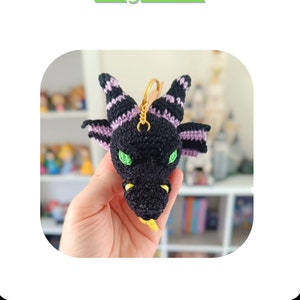 Puede incluir: Un llavero de drag&oacute;n de crochet negro y morado con ojos verdes y lengua amarilla. El llavero est&aacute; colgado de una cadena dorada.