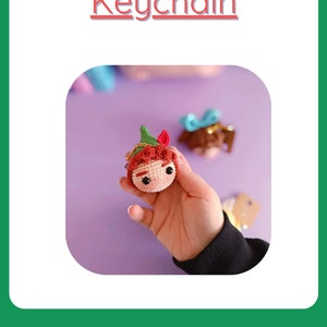 May include: Crocheted Peter Pan keychain with a green hat, red hair, and brown eyes. The keychain is held in a hand against a purple background.
