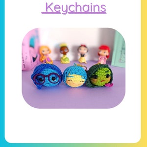 May include: Three crocheted keychains of the characters Joy, Sadness, and Disgust from the movie Inside Out. The keychains are on a purple surface. The text "Joy, Sadness, Disgust Keychains" is at the top of the image.