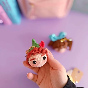 Puede incluir: Un personaje de crochet con pelo rojo, un sombrero verde y ojos marrones. El personaje sostiene una cadena dorada en su mano.