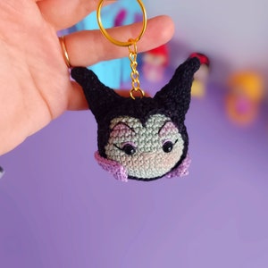 Puede incluir: Un llavero de Maléfica de crochet morado y negro con una cadena y un anillo dorados. El llavero tiene la forma de la cabeza de Maléfica con ojos grandes y un lazo morado.